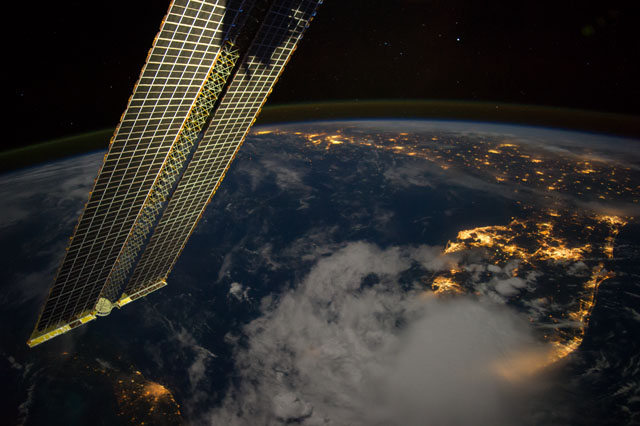 image at earth's horizon containing a solar panel, bright lights from cities, and a large weather pattern